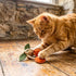 A playful orange tabby cat interacts with a cherry-themed pet toy featuring green leaves and orange fruit. The cat is positioned on a wooden floor, showcasing its curiosity as it paws at the toy in a sunlit room.