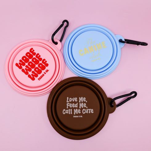 Three collapsible silicone dog bowls are displayed on a pastel pink background. The bowls are in pink, blue, and brown, featuring playful text designs that read Woof Woof Woof, The Canine Club, and Love Me, Feed Me, Call Me Cute, each.