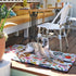 A greyhound rests on a colorful travel mat adorned with floral patterns, situated on a wooden patio. Surrounding the dog are wicker chairs and a table, with potted plants enhancing the vibrant outdoor setting.