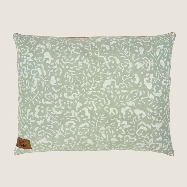 A rectangular dog bed featuring a soft sage green fabric adorned with a white floral and leaf pattern. The bed has a small tag in the lower corner that reads Indie Boho, emphasizing its stylish and cozy design.