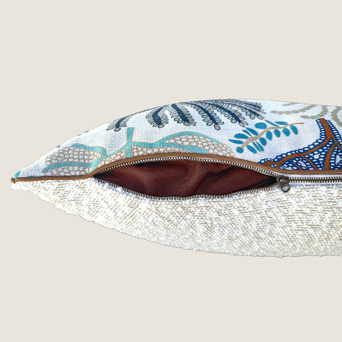 A close-up view of a decorative pillow featuring a colorful, patterned fabric with intricate designs in blue and orange, unzipped to reveal a soft, brown lining. The pillows texture contrasts with a smooth, white bouclé fabric.