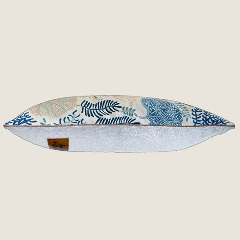 A decorative pillow featuring a cream-colored fabric with intricate botanical patterns in shades of blue and green. The pillow has a textured white bottom, adding depth and contrast to its design.