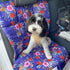 A fluffy black and white dog sits on a vibrant floral-patterned travel mat inside a car. The mat features a colorful design with pink and red flowers against a deep blue background, adding a cheerful touch to the vehicles interior.