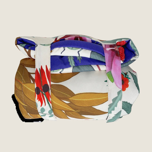 A rolled-up travel mat featuring vibrant illustrations of Australian wildflowers and plants, secured with white straps. The mat displays a colorful design on a pale yellow background, showcasing a blend of reds, greens, and blues.