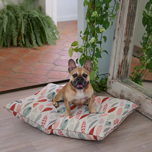 A tan French Bulldog sits comfortably on a colorful, leaf-patterned dog bed with shades of red and green. The background features a large mirror and lush greenery, creating a cozy indoor setting.