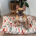 A tan French Bulldog sits happily on a stylish dog bed featuring a colorful leaf pattern in shades of red and green. In the background, a potted plant adds a touch of greenery to the modern, cozy setting.