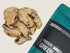 A pile of freeze-dried green lip mussels is displayed beside a teal and black packaging featuring the text 100% Freeze Dried Green Lip Mussels. The mussels have a natural, earthy appearance, highlighting their unique shape and texture.
