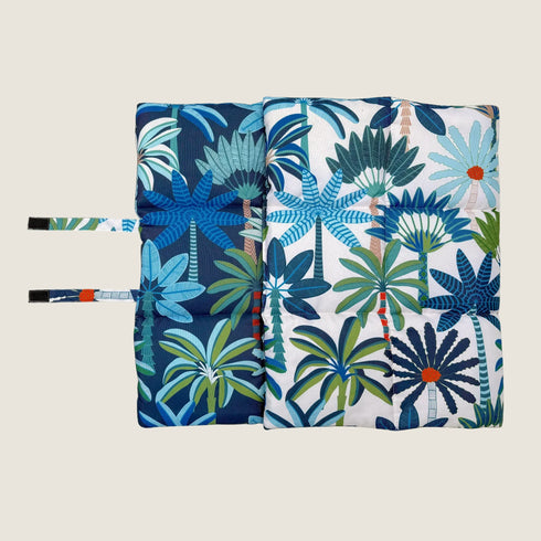 The image showcases a reversible dog travel mat featuring a vibrant coastal design. One side displays a bold pattern of tropical plants in shades of blue, green, and orange, while the other side features a contrasting design.