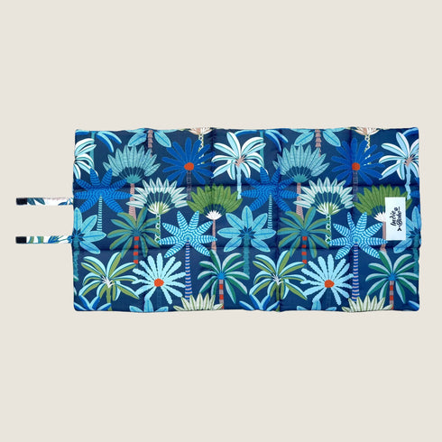 A vibrant dog travel mat featuring a bold coastal pattern with blue, green, and orange hues. The design showcases various stylized palm trees and tropical plants, emphasizing a lively and playful aesthetic.