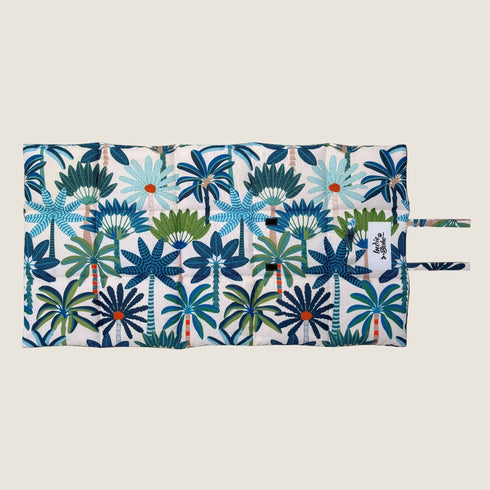 A colorful reversible travel mat featuring a vibrant coastal pattern with palm leaves in shades of blue, green, and orange. The mat has a quilted design and includes two fabric loops for easy storage or attachment.