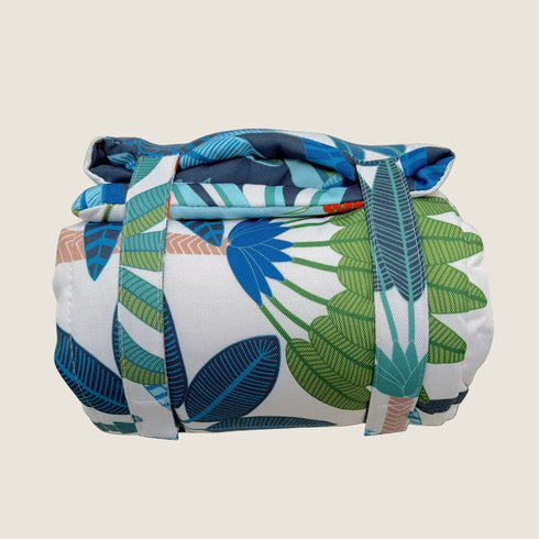 A rolled-up travel mat featuring a vibrant design with large green and blue leaves on a white background. The mat is secured with blue straps, showcasing its portability and stylish, tropical aesthetic.
