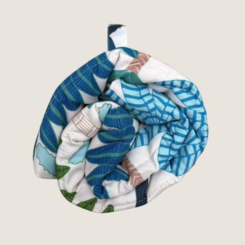A rolled-up dog travel mat features a vibrant design with a mix of blue and green coastal patterns, accented with touches of orange.