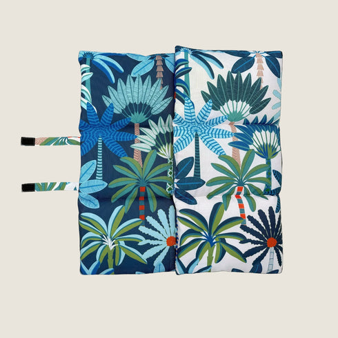 A reversible dog travel mat featuring a bold coastal design with vibrant blue, green, and orange palm tree patterns on one side and a contrasting pattern on the other. The mat is folded and secured with straps for easy transport.