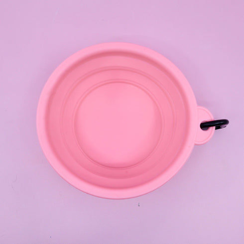 A pink collapsible silicone dog water bowl is displayed against a light purple background. The bowl features a clip for easy attachment, showcasing its practical design for travel and outdoor adventures.