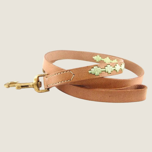 A handcrafted leather dog lead in a soft tan color features decorative embroidery with a light green pattern. The lead is complemented by a brass clip, showcasing a blend of natural materials and artisan craftsmanship.