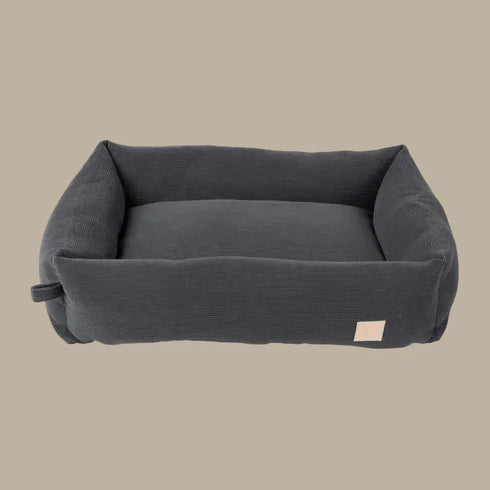 A plush, slate grey corduroy dog bed features high padded sides and a soft interior, designed for comfort. The bed has a simple, elegant shape and a small label on one side.