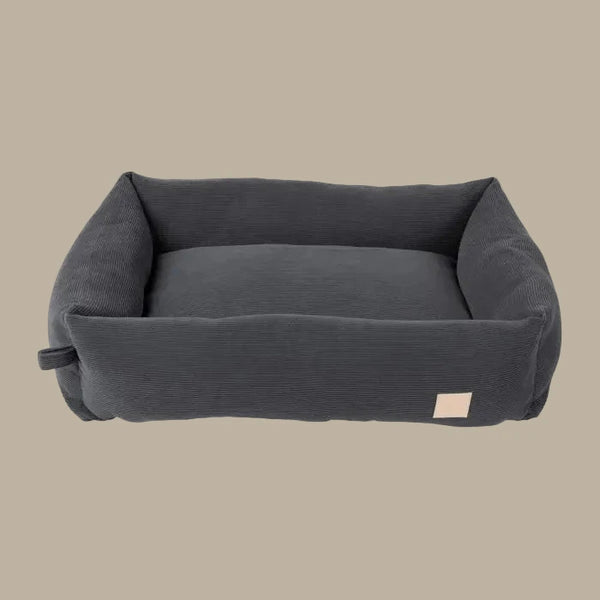 A plush, slate grey corduroy dog bed features high padded sides and a soft interior, designed for comfort. The bed has a simple, elegant shape and a small label on one side.