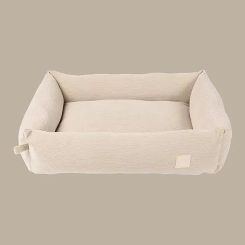 A plush, beige corduroy pet bed with a rectangular shape and softly padded sides, designed for comfort and style. The bed features a smooth interior and a subtle logo tag on one side.
