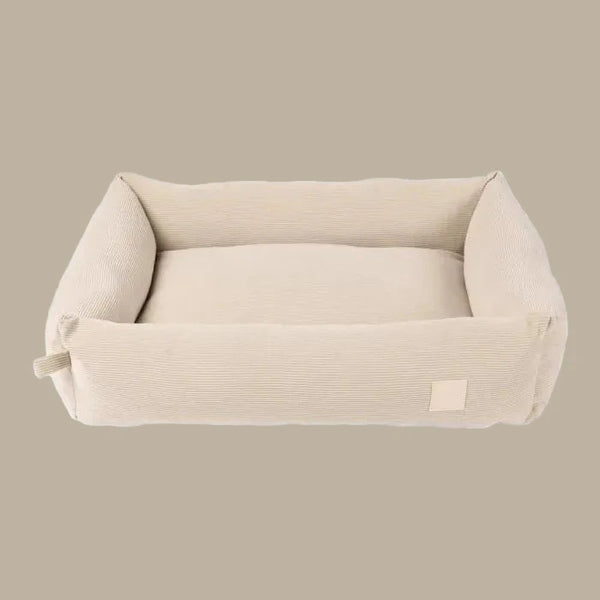 A plush, beige corduroy pet bed with a rectangular shape and softly padded sides, designed for comfort and style. The bed features a smooth interior and a subtle logo tag on one side.