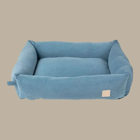 A soft, rectangular dog bed in a light French blue corduroy fabric features tall, cushioned sides and a plush center. The design includes a small tag on the side, blending style and comfort for pets.